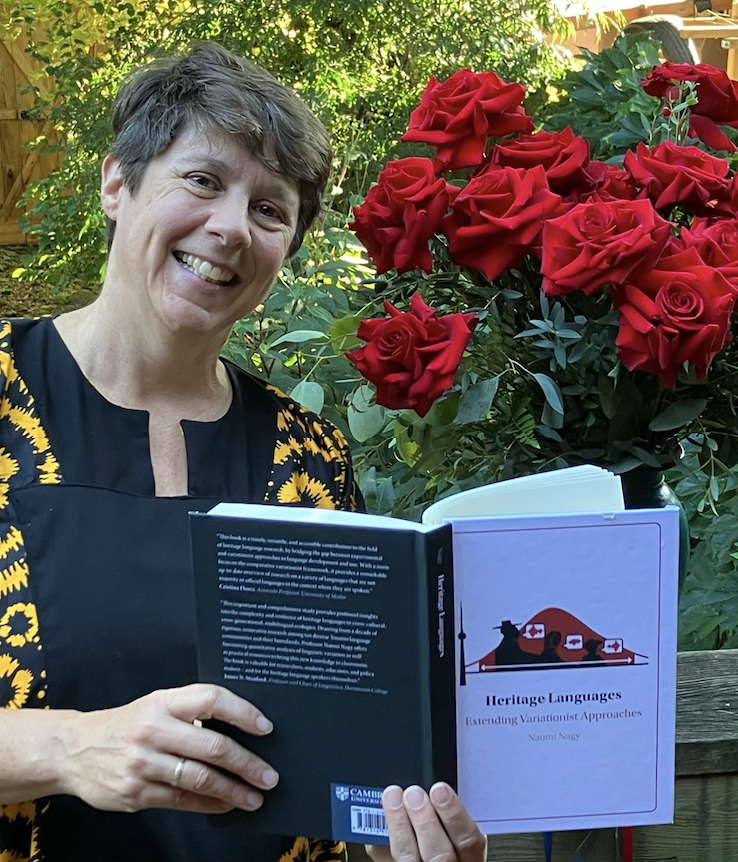 Professor Naomi Nagy, smiling, holding her new book in front of a flower bush